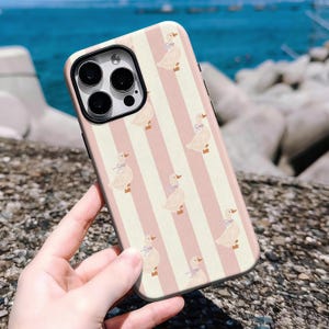 May include: A phone case featuring a repeating pattern of ducks with blue bows on a pink and white striped background. The case has a black camera lens frame and is held in a person's hand.