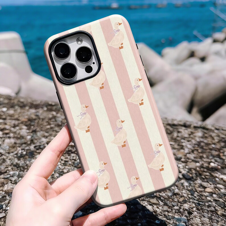 May include: A phone case featuring a repeating pattern of ducks with blue bows on a pink and white striped background. The case has a black camera lens frame and is held in a person's hand.