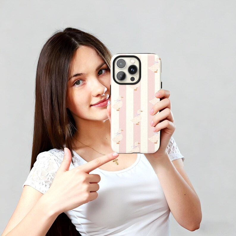 May include: A phone case featuring a pattern of white ducks on a pink and white striped background. The case has a black border and a camera cutout. The phone case is being held up by a person.