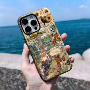 May include: A decorative phone case featuring a collage of sunflowers, a black cat, and floral and landscape imagery. The case includes the word "love" and other text elements. The case is held against a backdrop of the ocean.