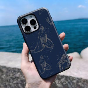 May include: Dark blue phone case with a gold whale design. The case has a pattern of whales and small star-like details. The phone case has a black border and a camera cutout. The background is a blurred view of the sea.