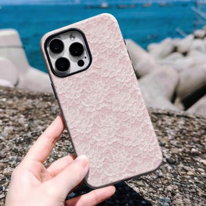 May include: A pink phone case with a white lace floral pattern. The case has a black camera lens frame and a black border. The phone case is held in a hand, with a blurred background of rocks and water.