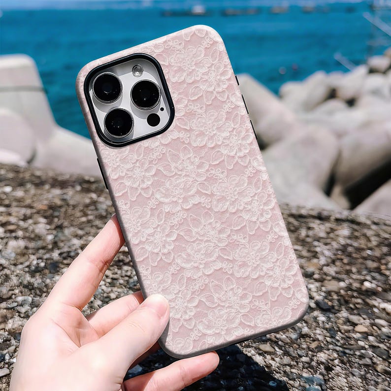 May include: A pink phone case with a white lace floral pattern. The case has a black camera lens frame and a black border. The phone case is held in a hand, with a blurred background of rocks and water.
