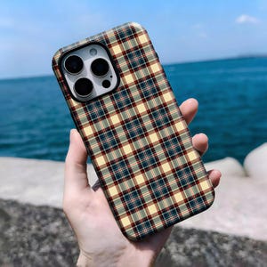 Puede incluir: Funda de teléfono con un diseño de cuadros en tonos beige, rojo, azul marino y gris. La funda tiene un acabado brillante y un recorte para la cámara. El fondo es una vista borrosa del mar y el cielo.