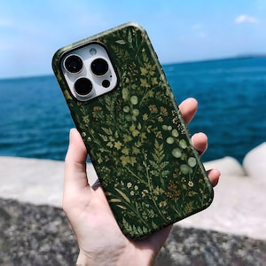 Op de afbeelding: Een donkergroen smartphonehoesje met een gedetailleerde botanische print van diverse wilde bloemen, varens en eucalyptusbladeren in tinten lichtgroen, geel en bruin. Het hoesje heeft een uitsparing voor een camera met drie lenzen aan de achterkant.