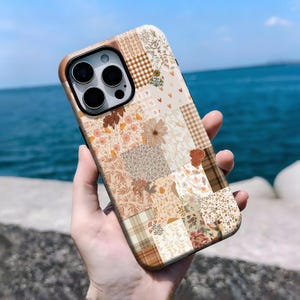 May include: A phone case with a patchwork design in shades of brown, beige, and cream. The case has a brown border and a camera cutout. The design includes floral, plaid, and leaf motifs, creating a vintage aesthetic. The case is held against a backdrop of the sea.