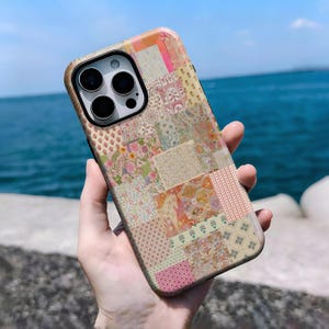May include: A colorful phone case with a patchwork design. The case features a variety of floral and geometric patterns in shades of pink, beige, and orange. The phone case has a black camera module.