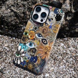 May include: A detailed, multi-coloured phone case featuring eye motifs, suns, and celestial elements. The case is embellished with beads and intricate patterns. It rests on a bed of small pebbles. The design is intricate and eye-catching.