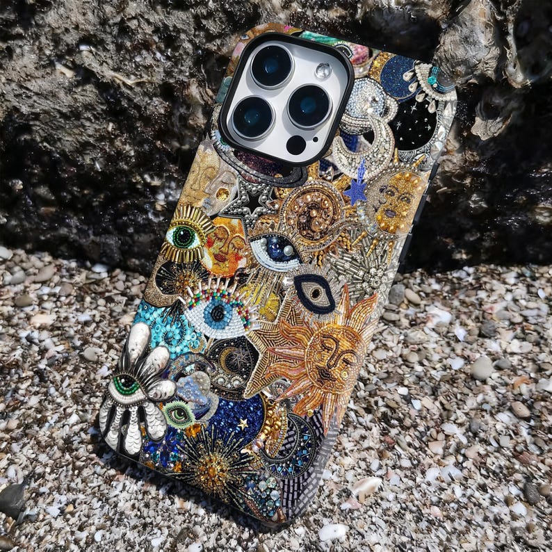 May include: A detailed, multi-coloured phone case featuring eye motifs, suns, and celestial elements. The case is embellished with beads and intricate patterns. It rests on a bed of small pebbles. The design is intricate and eye-catching.