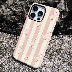 May include: A phone case with a pink and white striped pattern. The case features a repeating design of cartoon ducks wearing bows. The phone case is shown on a rocky surface.