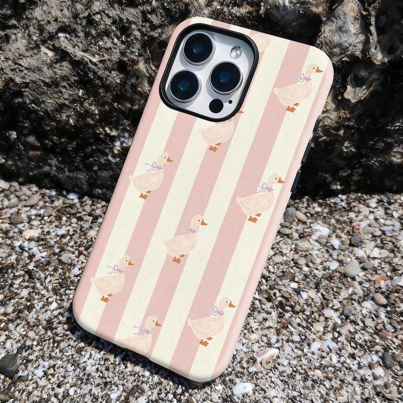 May include: A phone case with a pink and white striped pattern. The case features a repeating design of cartoon ducks wearing bows. The phone case is shown on a rocky surface.