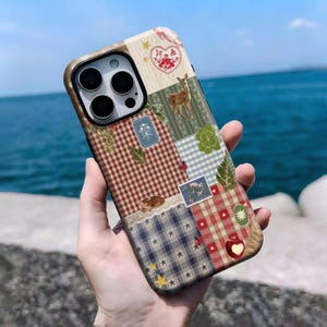 Puede incluir: Funda de teléfono de estilo patchwork con una variedad de patrones e ilustraciones. El diseño incluye cuadros rojos, cuadros azules, motivos florales y ciervos. También se ven un corazón, estrellas y una pequeña manzana. La funda se sostiene contra el mar.