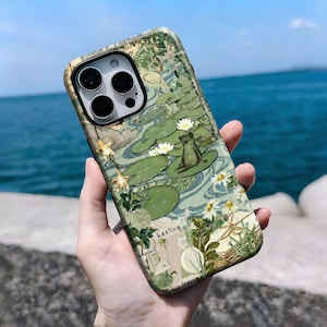 Puede incluir: Funda de teléfono con un diseño de estanque, con nenúfares, flores blancas y una rana. El diseño de estilo vintage incorpora elementos botánicos y el texto "healing happens in". La funda tiene un acabado brillante.