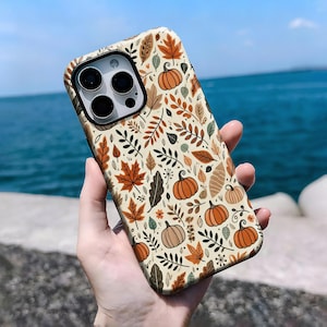 May include: A phone case with a fall-themed pattern. The case features a cream background with illustrations of pumpkins, leaves, and floral elements in shades of orange, brown, and green. The case is held in a person's hand.