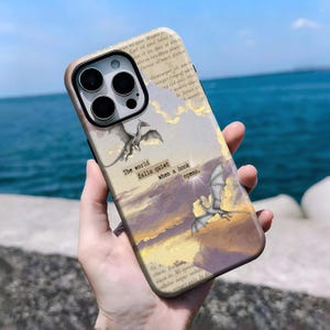 Könnte beinhalten: Eine Smartphone-Hülle mit einem Hintergrund aus einer alten Buchseite, zwei fliegenden Drachen, Goldfolien-Akzenten und dem aufgedruckten Text, The world falls quiet when a book opens. Das Design zeigt einen sonnenuntergangsfarbenen Himmel.