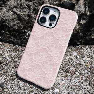 May include: A pink phone case with a white floral lace pattern. The case has a black border around the camera and a black frame. The phone case is lying on a surface of small pebbles and sand.