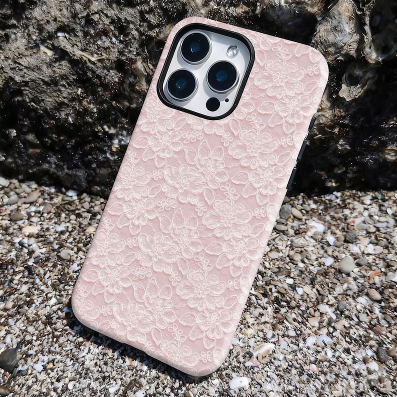 May include: A pink phone case with a white floral lace pattern. The case has a black border around the camera and a black frame. The phone case is lying on a surface of small pebbles and sand.