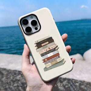 Könnte beinhalten: Cremefarbene Handyhülle mit einem Design aus gestapelten Büchern. Die Buchrücken zeigen Titel wie "Midnights" und "Reputation". Die Hülle hat ein schwarzes Kameramodul. Der Hintergrund ist das Meer.