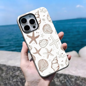 Puede incluir: Una funda de teléfono blanca con un patrón repetido de conchas marinas y estrellas de mar en tonos marrones. La funda tiene un módulo de cámara negro y se sostiene contra un fondo marino.