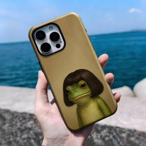 May include: A gold-colored phone case featuring a cartoon frog with a brown bob haircut. The frog is green and has large, expressive eyes. The case has a camera cutout and a smooth finish.