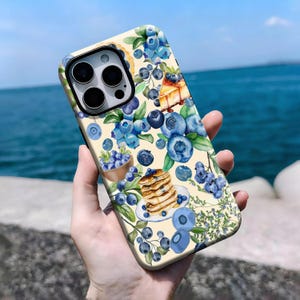 May include: A phone case featuring a watercolor design of blueberries, pancakes, and pie slices on a cream-colored background. The case has a glossy finish and a black camera module.