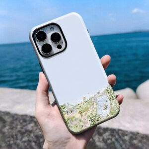 May include: A white phone case with a black camera module and a watercolour illustration of a park scene. The case features a detailed design of a park with people, trees, and a body of water.