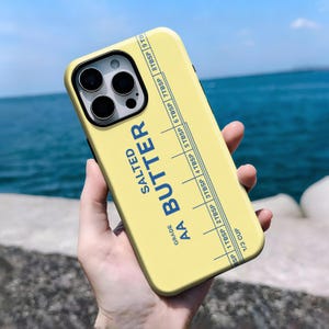 May include: A yellow phone case with a butter-themed design. The case features the words "SALTED BUTTER" and a measuring guide in blue. The case also has the text "AA GRADE" and a measurement scale.