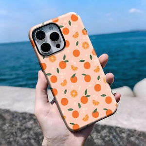 May include: A peach-colored phone case featuring a repeating pattern of oranges, orange slices, green leaves, and small white flowers. The case has a black camera lens cutout and a glossy finish. The phone case is held in a hand.