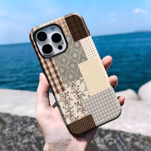 Può includere: Una custodia per smartphone con un design patchwork di vari motivi marroni, beige e crema. La superficie mostra un mix di quadri, fiori, pois e stampe botaniche disposti in una griglia, con un'apertura per la fotocamera a tre obiettivi.