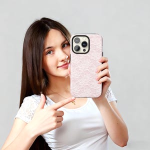 May include: A light pink phone case with a delicate lace pattern. The case has a black border and a camera cutout. The phone case is held up by a person.