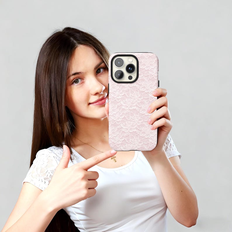 May include: A light pink phone case with a delicate lace pattern. The case has a black border and a camera cutout. The phone case is held up by a person.