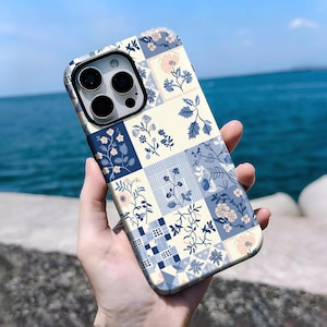 Pode incluir: Uma capa de telefone com um design de patchwork floral em azul, creme e rosa. A capa apresenta um padrão quadrado com vários motivos florais e de folhas. A capa do telefone é segurada contra um fundo marinho.