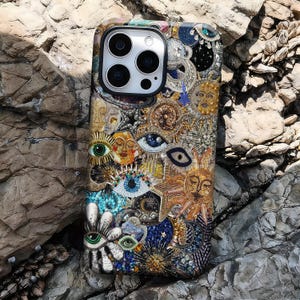 May include: A detailed phone case with a celestial and mystical design. The case features suns, moons, stars, and eyes, with gold, blue, and white beadwork. The case is designed to fit a smartphone, offering both protection and a unique aesthetic.