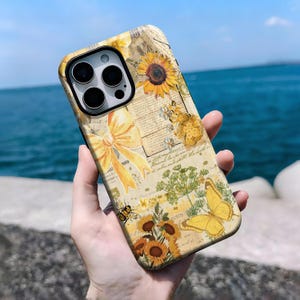 May include: A decorative phone case featuring a vintage design with sunflowers, butterflies, and a bee. The case has a yellow bow and text on a cream-colored background. The case is held in a hand.