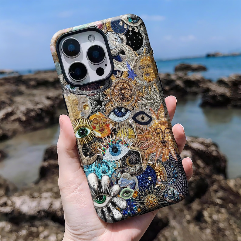 May include: A detailed phone case with a celestial design. The case features suns, moons, and eyes in gold, blue, and white. The case is held against a backdrop of rocks and the sea.