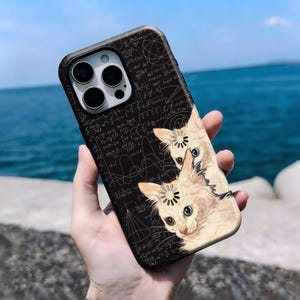 Op de afbeelding: Een zwarte smartphonehoes met een wit patroon van wiskundige formules en twee lichtgekleurde kittens. Elk kitten heeft een cirkelvormig laadicoon op het voorhoofd. De hoes heeft een uitsparing voor een camerasysteem met drie lenzen.