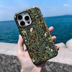 May include: A phone case with a dark background and a botanical pattern featuring green ferns, white flowers, and brown accents. The case has a camera cutout with three lenses.