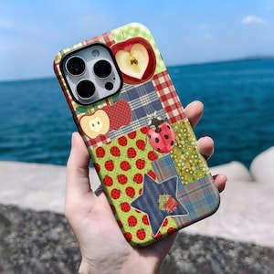 Può includere: Una custodia per telefono colorata con disegni di mele e coccinelle. La custodia presenta motivi a quadri, a pois e floreali. È visibile una mela rossa con un cuore. La custodia è tenuta in mano.