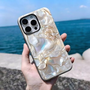 Può includere: Una custodia per telefono perlata con un design marmorizzato a spirale nei toni del bianco, crema e oro. La custodia ha un ritaglio nero per la fotocamera ed è tenuta su uno sfondo di mare e cielo.