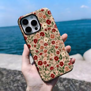 May include: A floral phone case featuring a pattern of red, white, and pink flowers with green leaves on a tan background. The case has a black camera module and is held against a backdrop of the ocean and sky.