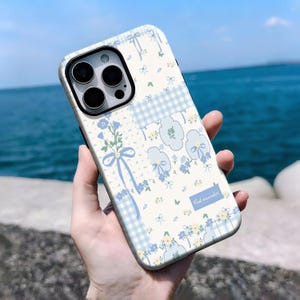 Pode incluir: Capa de telefone floral branca e azul com um padrão xadrez vichy azul claro. O design apresenta flores azuis, laços e o texto "first encounter". A capa do telefone está em uma mão.