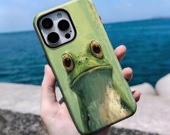 以下が含まれることがあります： カエルのイラストが描かれた緑色の携帯電話ケース。ケースはマット仕上げで、大きく表現力豊かな目を持つカエルの詳細な絵が描かれています。ケースは海の背景に対して保持されています。