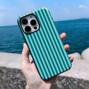 Puede incluir: Funda de teléfono a rayas turquesa y verde azulado. La funda tiene un borde negro para la lente de la cámara y está sujeta en la mano de una persona. El fondo muestra el mar y el cielo.
