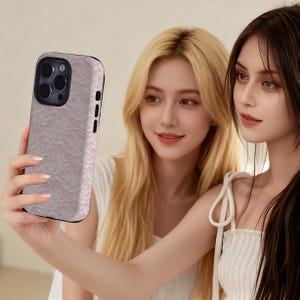 May include: A smartphone case with a pink lace pattern. The case has a black border and a camera cutout. The phone case is being held by a person.