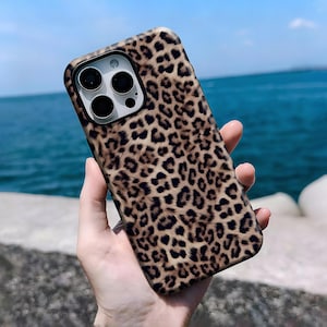 May include: A phone case with a leopard print design. The case is brown and tan with black spots. The phone case has a camera cutout with three lenses and a flash. The background is a blurred view of the ocean and sky.