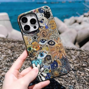 May include: A detailed phone case with a mosaic of suns, eyes, and celestial motifs in gold, blue, and white. The case has a textured finish and a black camera lens aperture.