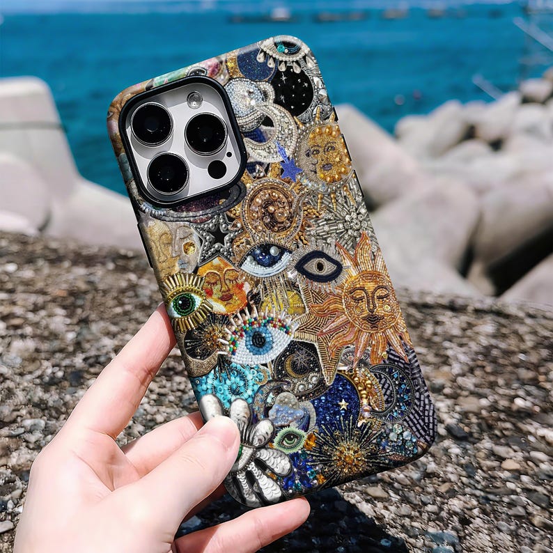 May include: A detailed phone case with a mosaic of suns, eyes, and celestial motifs in gold, blue, and white. The case has a textured finish and a black camera lens aperture.