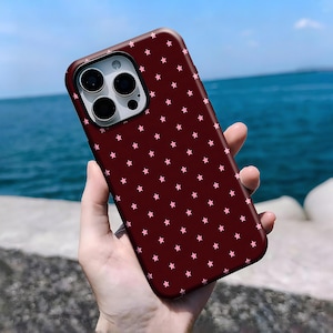 May include: A burgundy phone case with a pattern of small pink stars. The case has a smooth finish and is designed to fit a smartphone with multiple camera lenses.