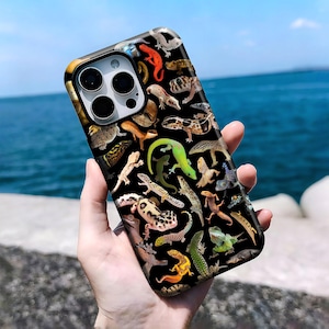 Op de afbeelding: Een zwarte smartphonehoes met een dicht, kleurrijk patroon van verschillende hagedissoorten, waaronder gekko's en kameleons, in tinten groen, oranje, bruin en geel. De hoes heeft een uitsparing voor een drievoudig camerasysteem aan de achterkant.