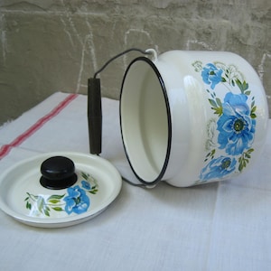 May include: A white enamel canister with a black rim and a matching lid. The canister features a blue floral pattern with green leaves. It includes a metal swing handle with a dark brown cylindrical grip and a black knob on the lid.